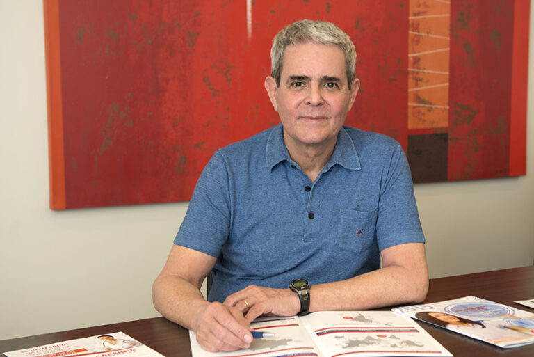 Paulo Milliet Roque, um homem de cabelos grisalhos, está sentado, vestindo uma camisa polo azul, um relógio preto e na frente de um quadro laranja e vermelho.